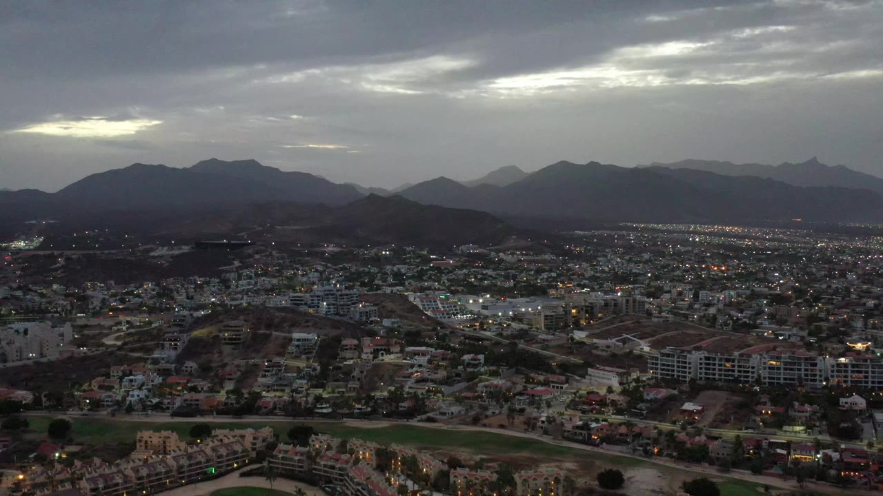 Los Cabos aerial view one second in, showing clustered buildings and roads beneath broad mountain silhouettes and a dim cloud break, with no visible people.