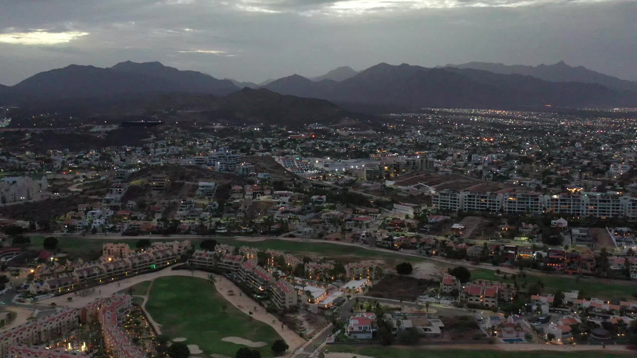 Aerial Los Cabos cityscape showing residential blocks, road grids, a green course edge near the bottom, distant lights, layered mountains, cloudy sky, and no visible people.
