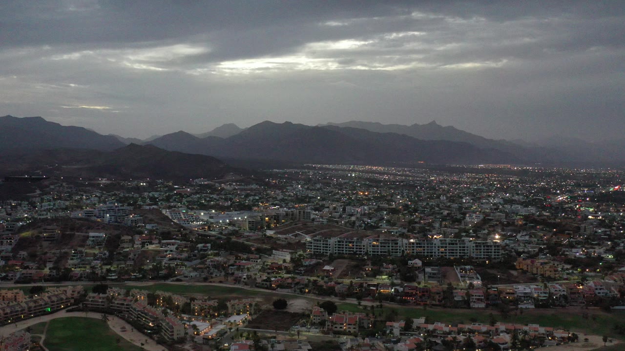 Wide aerial view over Los Cabos, Mexico, with city lights below, dark mountain silhouettes on the horizon, layered gray clouds above, and no visible people.