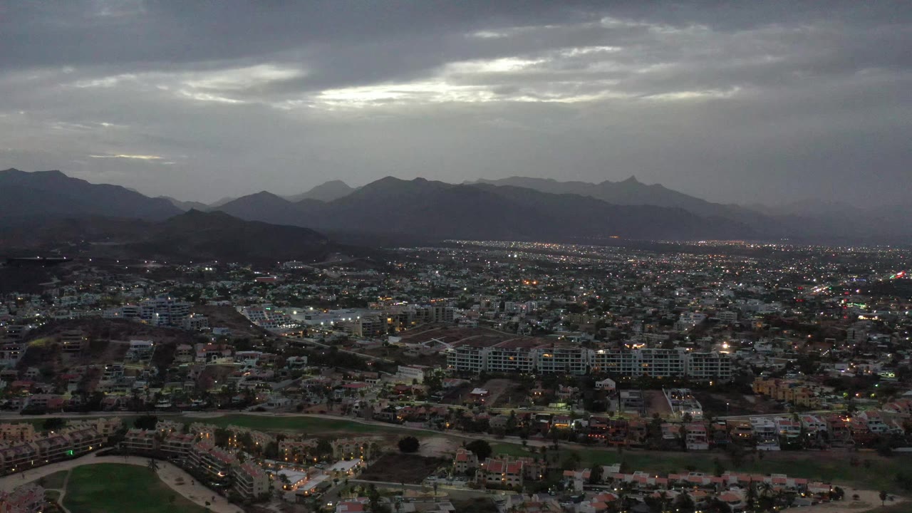 Aerial Los Cabos cityscape with illuminated blocks across the lower frame, a dark ridge line in the background, bright cloud breaks overhead, and no visible people.