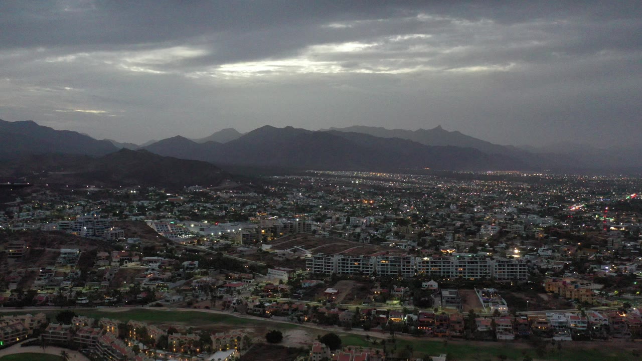 Wide aerial view of Los Cabos, Mexico at dusk, with city blocks and road lights below layered mountain silhouettes and no visible people.