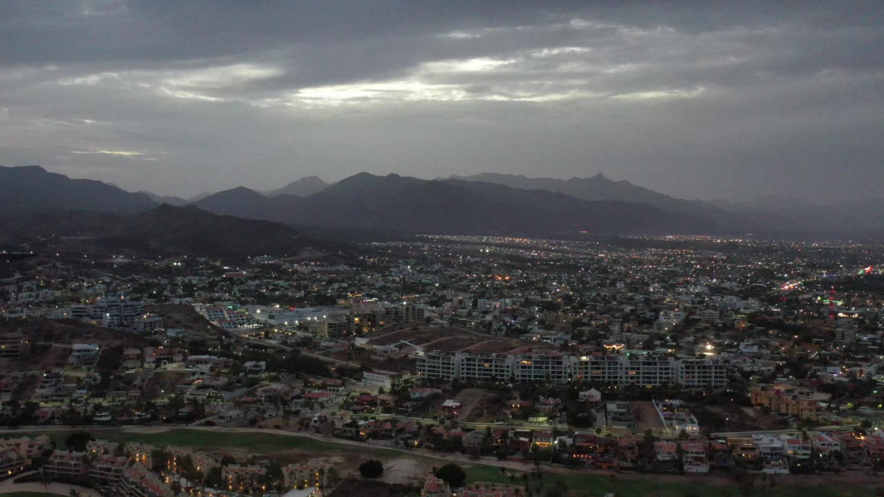 A wide Los Cabos panorama shows scattered city lights, low-rise buildings, and jagged mountain silhouettes below a lighter opening in the cloud cover, with no visible people.