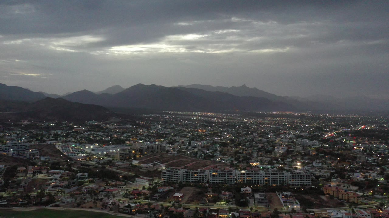 Wide aerial view of a city below dark clouds and distant mountains.