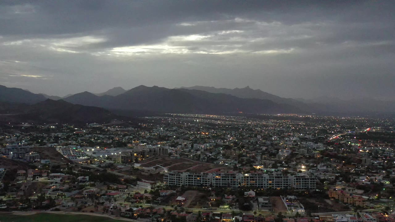 A broad Los Cabos aerial view shows residential and commercial blocks across the lower frame, layered mountains in the distance, dim city lights, and no visible people.