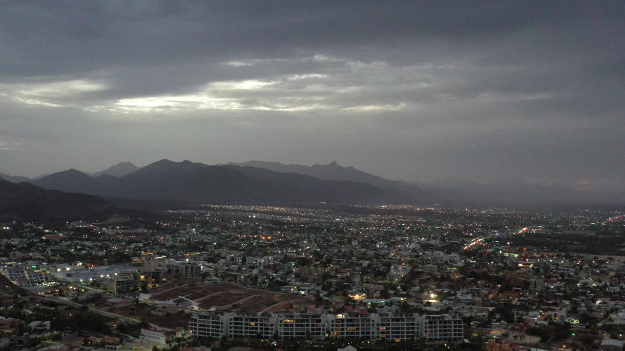 Wide aerial view over Los Cabos, Mexico, with scattered city lights, dark mountain silhouettes on the horizon, layered gray clouds, and no visible people.