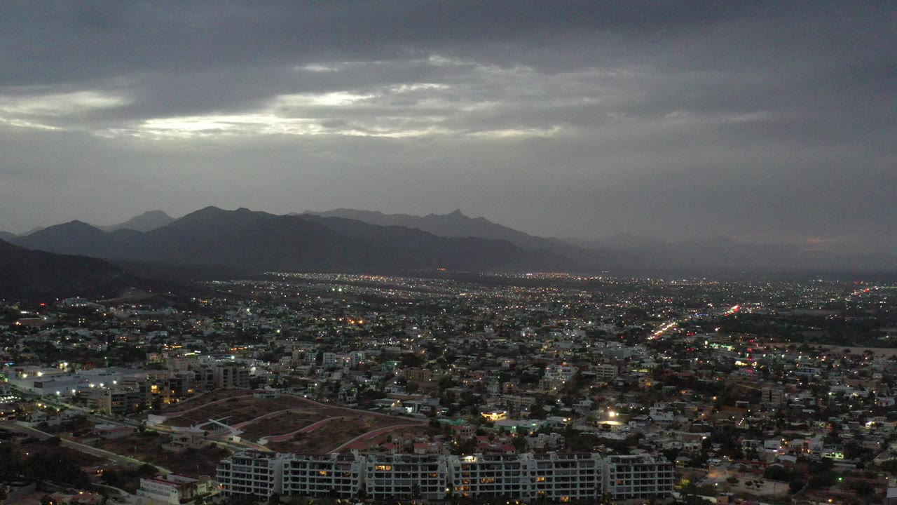 Wide aerial view of a city below dark clouds and distant mountains.