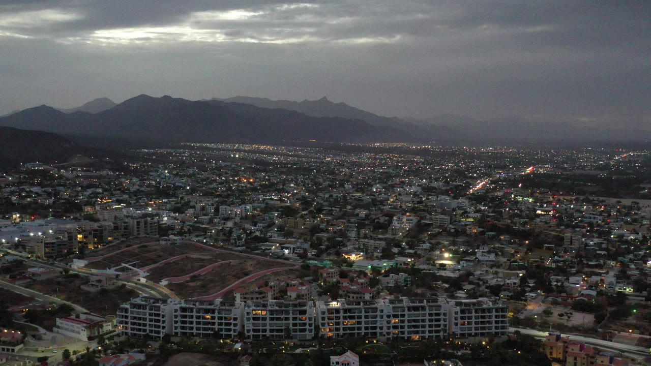 Wide aerial view of a city below dark clouds and mountains.