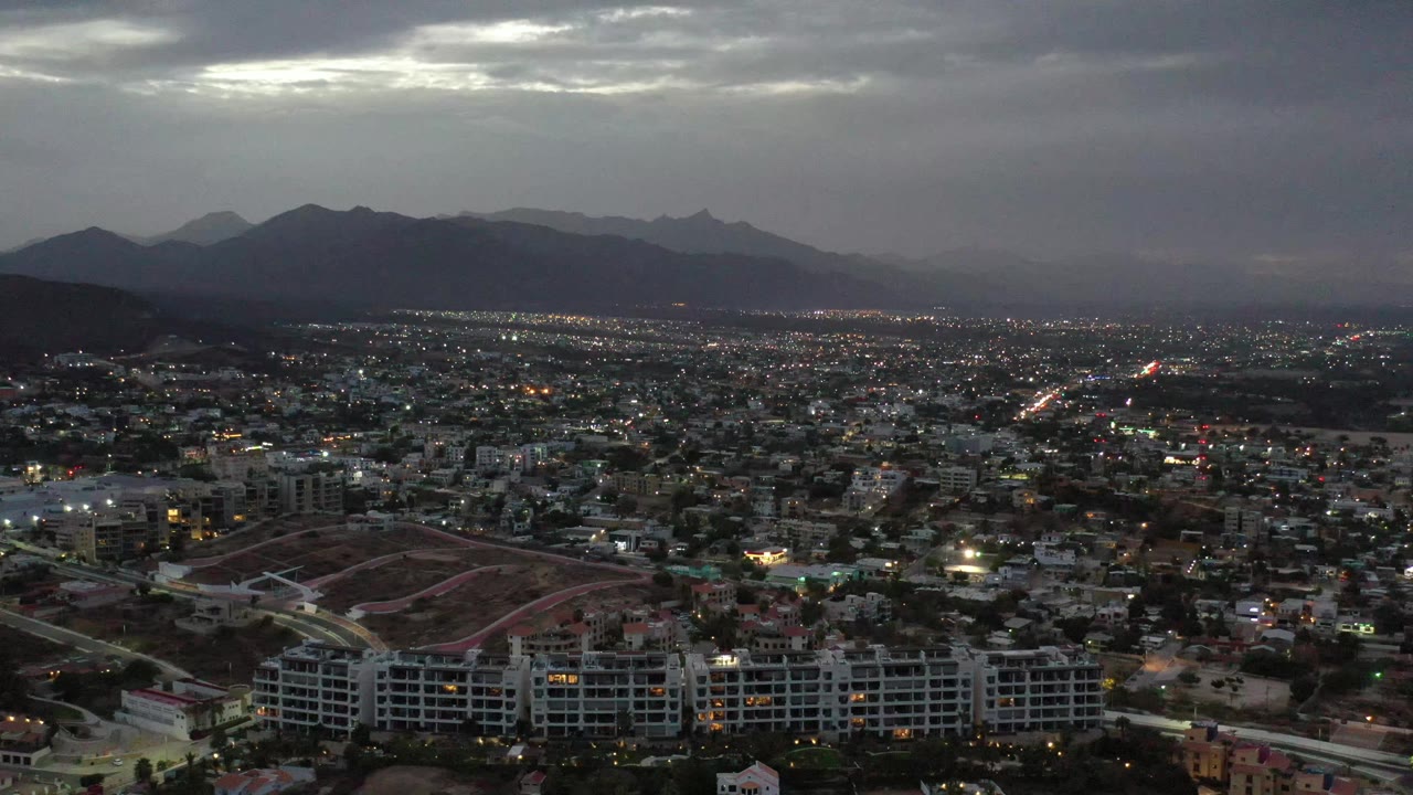 Los Cabos urban blocks and curving roads sit below a layered mountain backdrop, with a bright break in the overcast sky and no visible people.