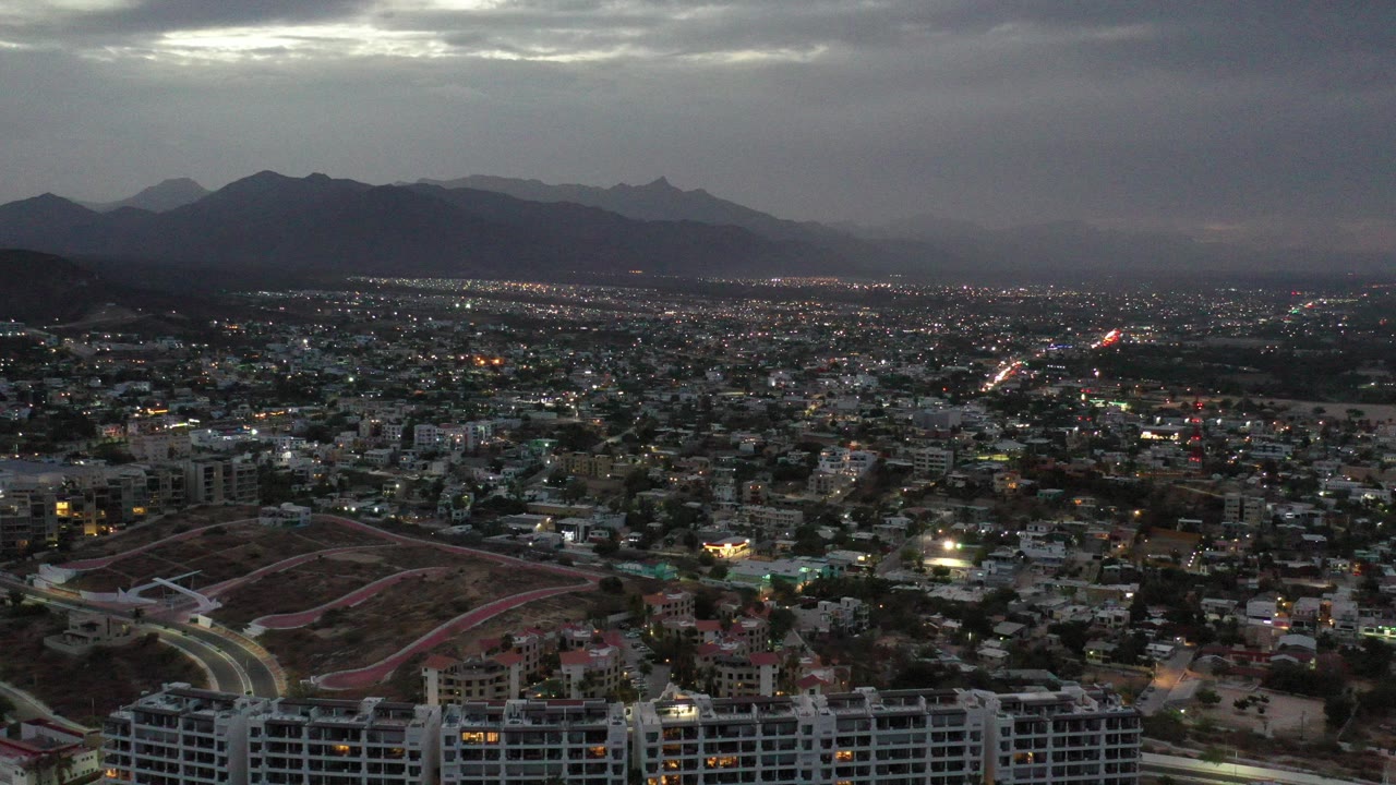 Los Cabos, Mexico aerial city view at dusk, with hillside roads and rooftops in the foreground, dark mountains in the distance, gray cloud cover, and no visible people.