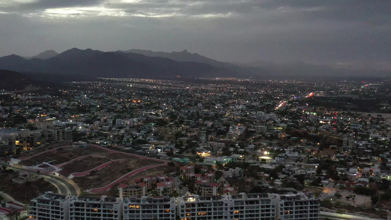 Los Cabos, Mexico city lights spread across the frame beneath a layered mountain skyline and low gray evening clouds, with no visible people.