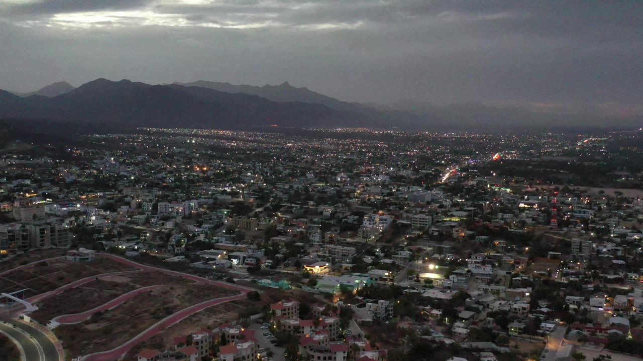 Elevated view across Los Cabos, Mexico, showing a foreground reddish hillside with pale roads, clustered buildings, distant mountains, gray sky, and no visible people.