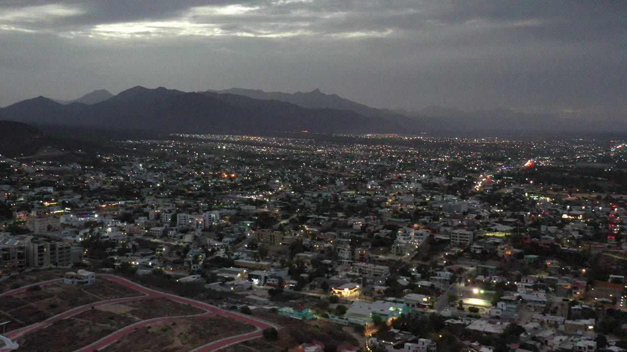Wide elevated view over Los Cabos at dusk, with city buildings and street lights below, dark mountains on the horizon, heavy clouds overhead, and no visible people.