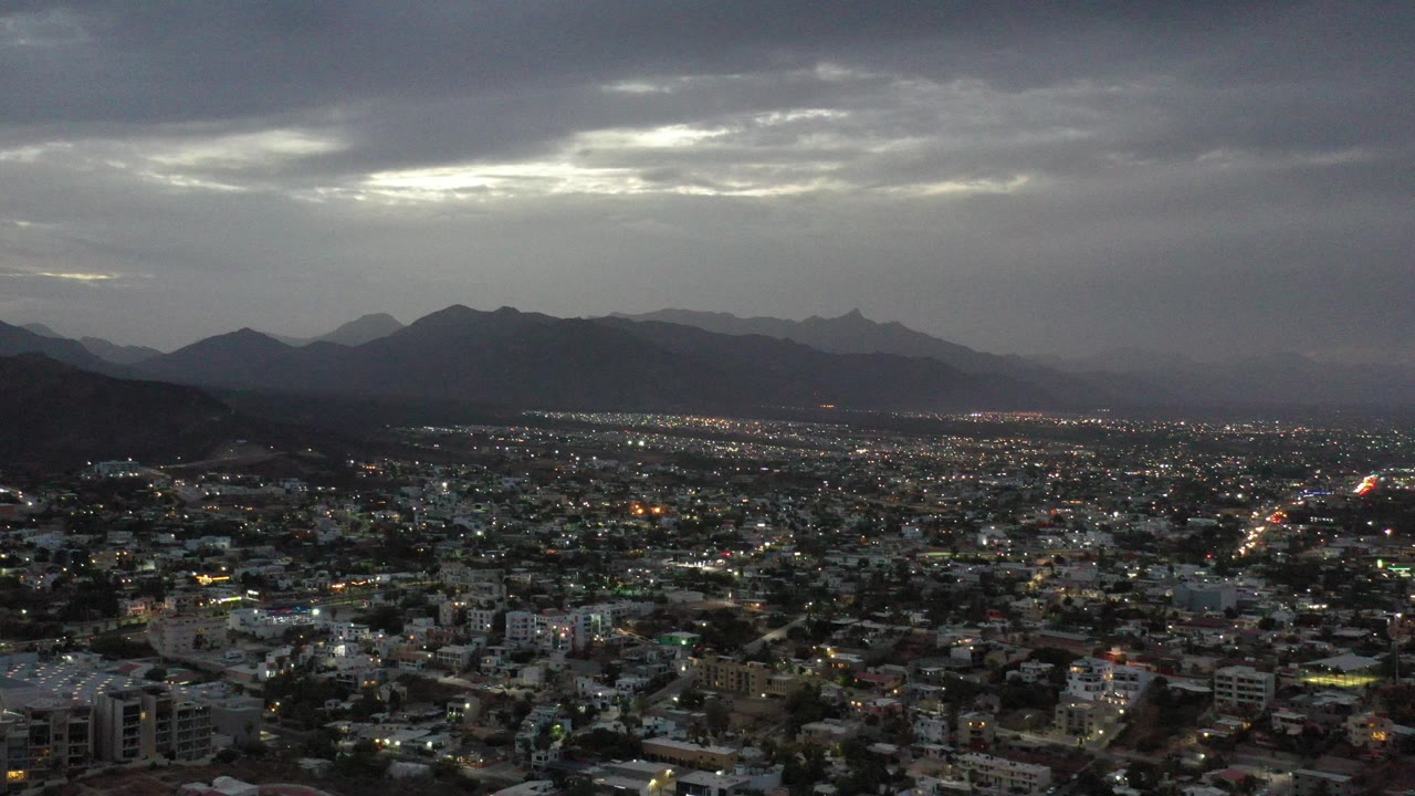 Wide aerial view of a city below dark mountains and cloudy sky.