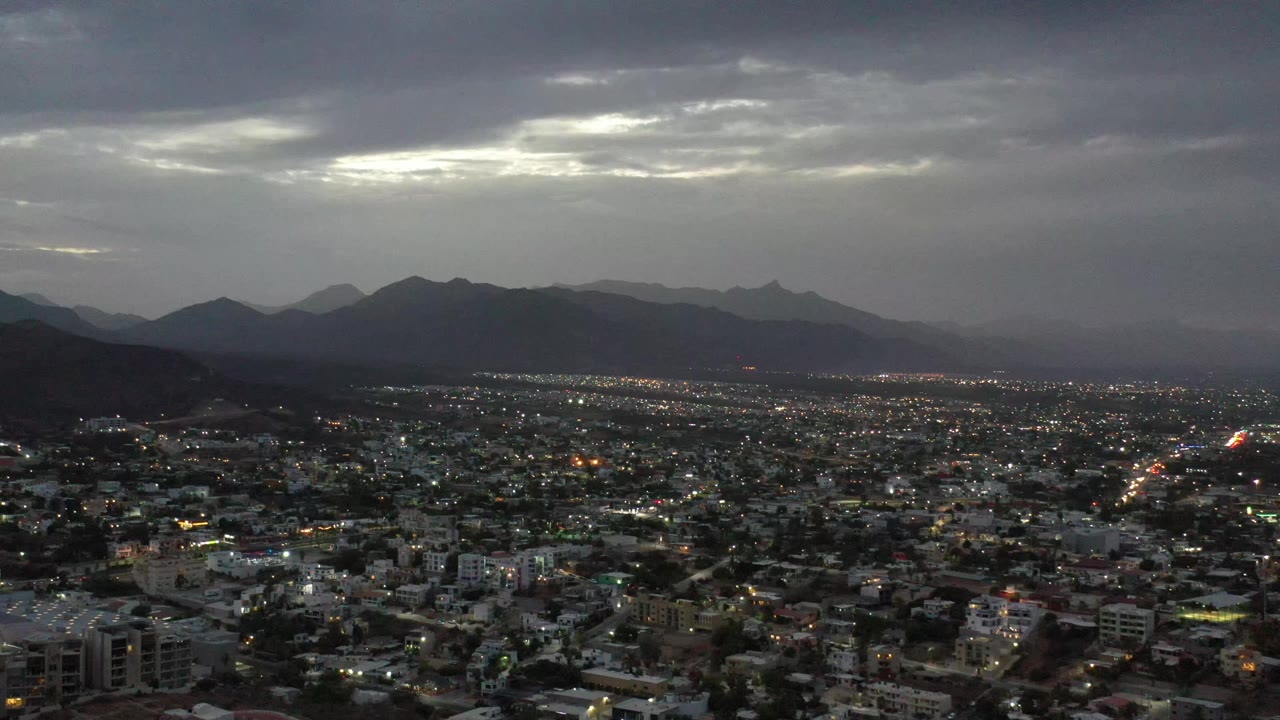 Elevated Los Cabos view showing a lit urban grid across the lower frame, layered mountains in the distance, a pale break in the clouds, and no visible people.