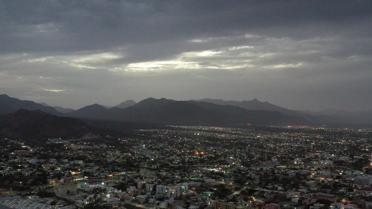 Wide elevated view over Los Cabos, Mexico, with city lights below, dark mountain silhouettes on the horizon, pale sunlight through heavy clouds, and no visible people.