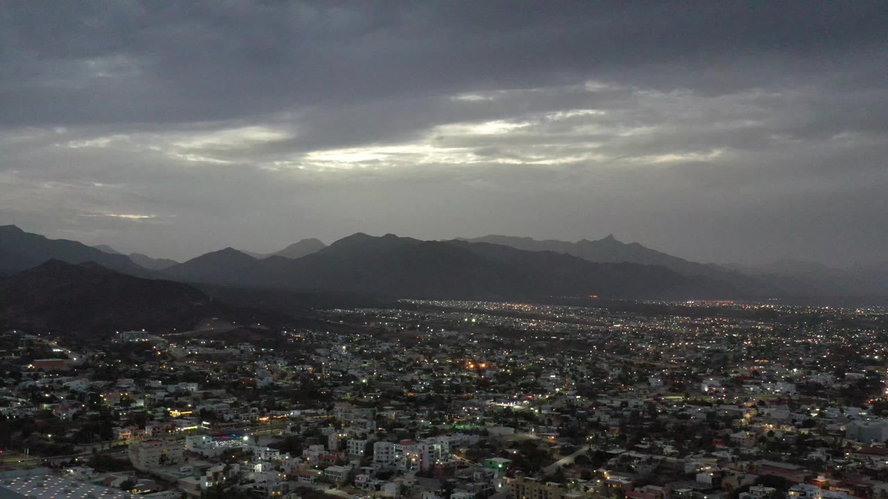 Los Cabos, Mexico appears from a high vantage point with bright city lights in the lower frame, layered mountain ridges in the distance, gray clouds overhead, and no visible people.