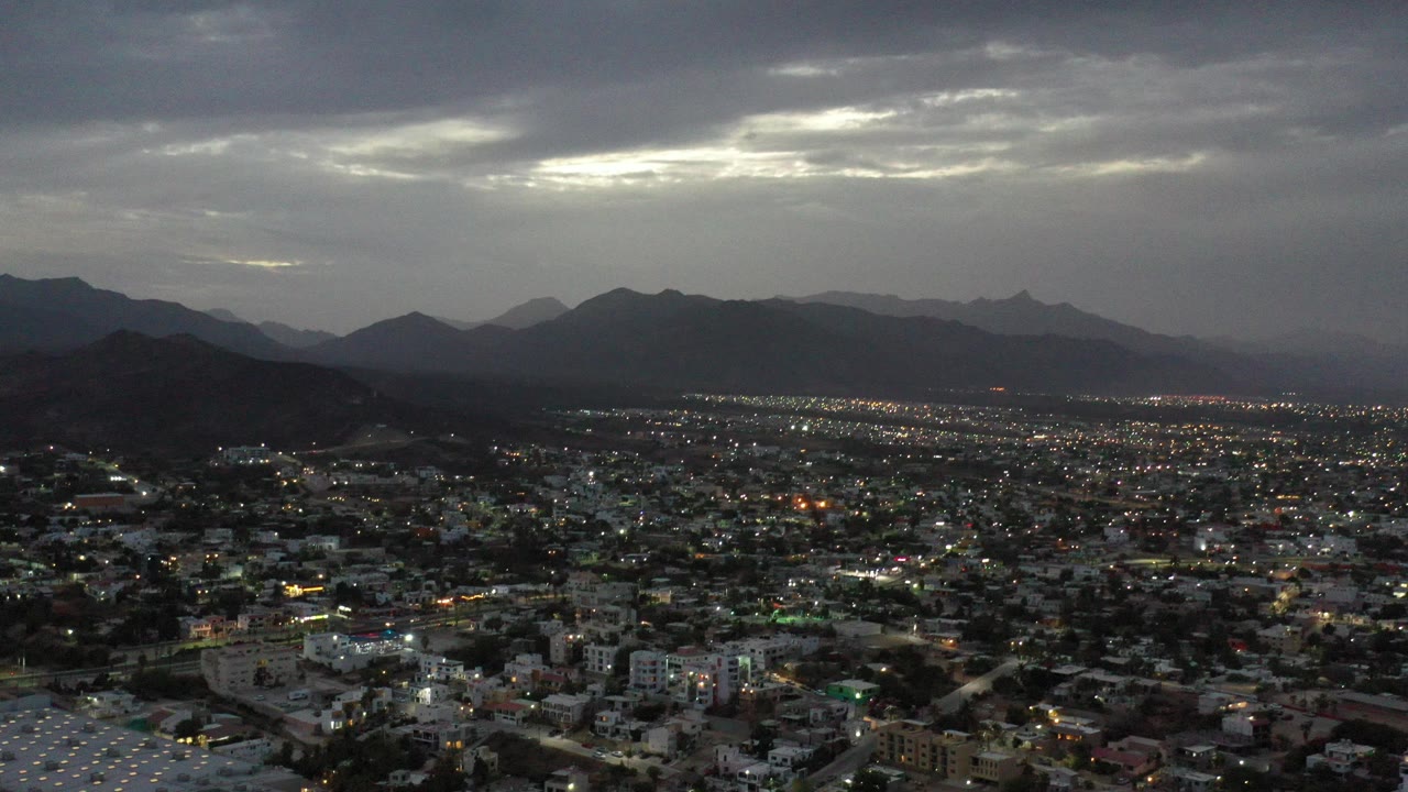 Opening aerial view over Los Cabos, Mexico, with scattered city lights, dark mountain silhouettes, a gray cloud ceiling, and no visible people.
