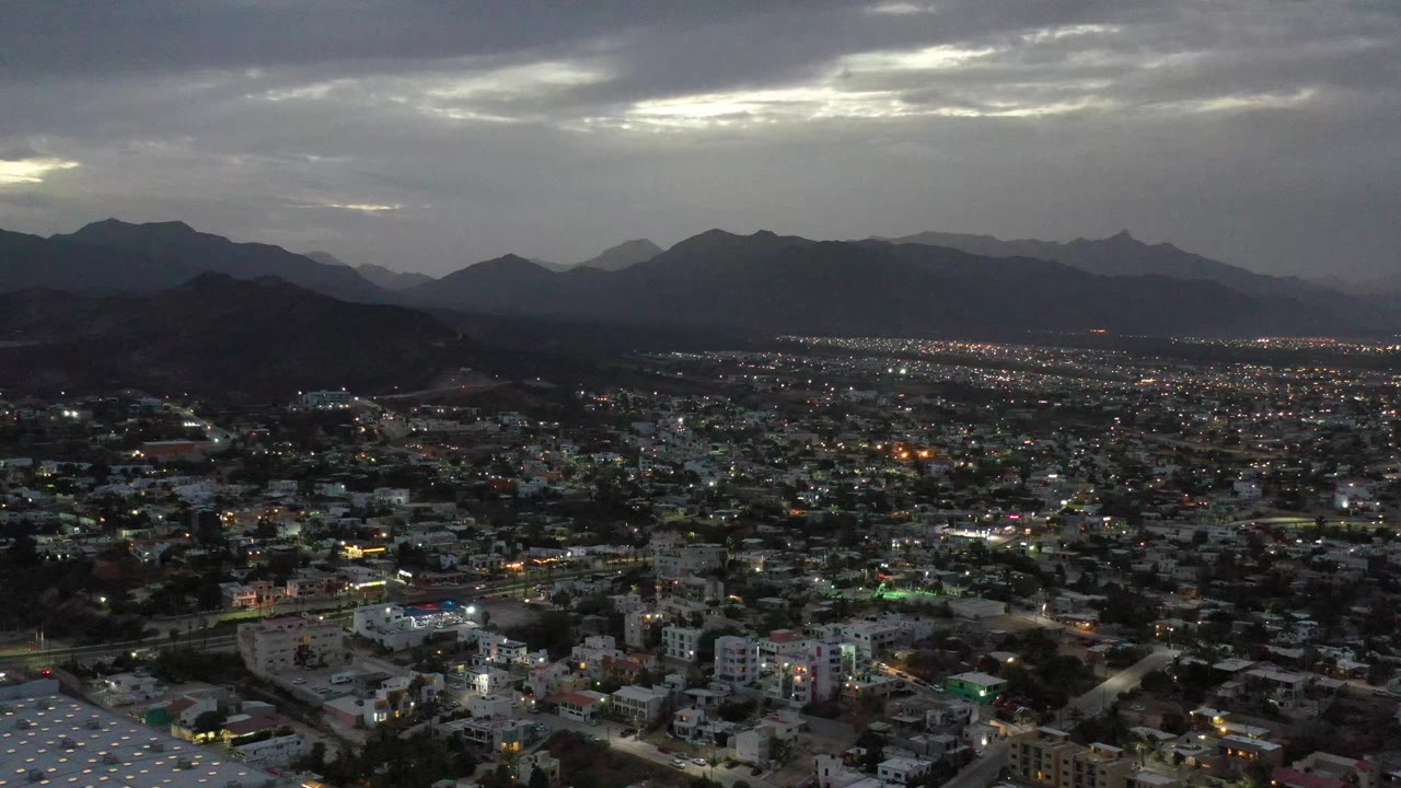 Los Cabos city lights begin to show across the urban grid beneath dark hills and layered gray clouds, with no visible people.