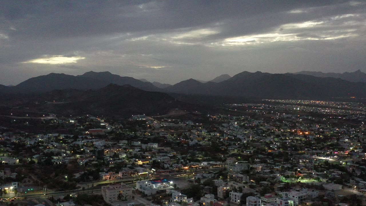 Opening frame of Los Cabos, Mexico at night, with illuminated streets and buildings below dark mountain ridges, layered clouds, and no visible people.