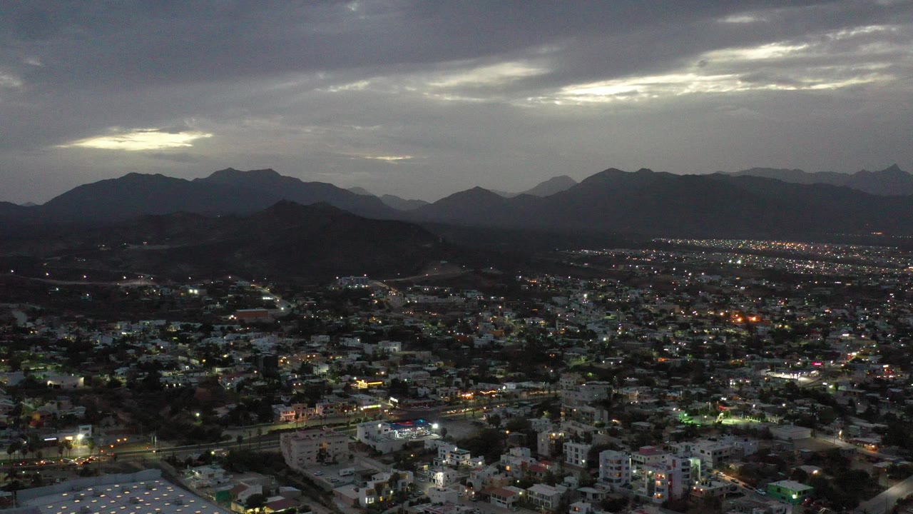 Wide aerial view over Los Cabos, Mexico at dusk, with city lights beginning to glow beneath dark mountain silhouettes and a gray cloud layer, no visible people.
