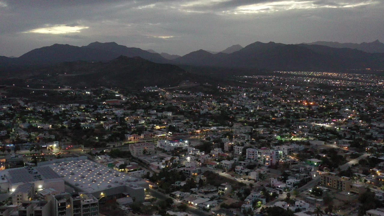 Wide aerial view over Los Cabos, Mexico, with scattered town lights, low buildings, a white-roofed structure in the foreground, dark mountain ridges, cloudy sky, and no visible people.
