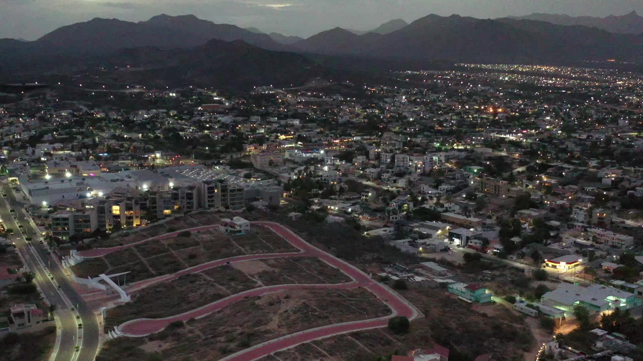 Los Cabos appears from a high aerial angle with a bright road corridor at left, scattered building lights across the midground, dark mountains behind the city, and no visible people.