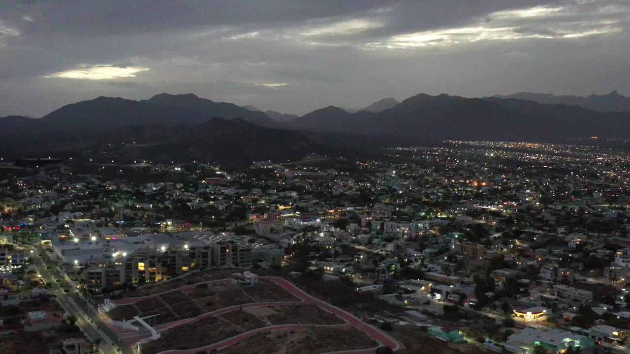 Los Cabos aerial view at dusk with more city lights visible across the middle distance, a dark ridge line, pale openings in the cloud cover, and no visible people.