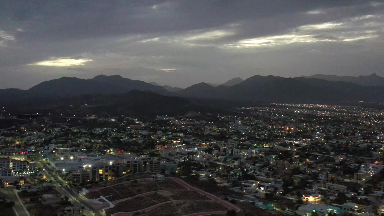 A high drone view of Los Cabos shows more of the illuminated town grid beneath layered mountain ridges and a pale break in the cloudy sky, with no visible people.