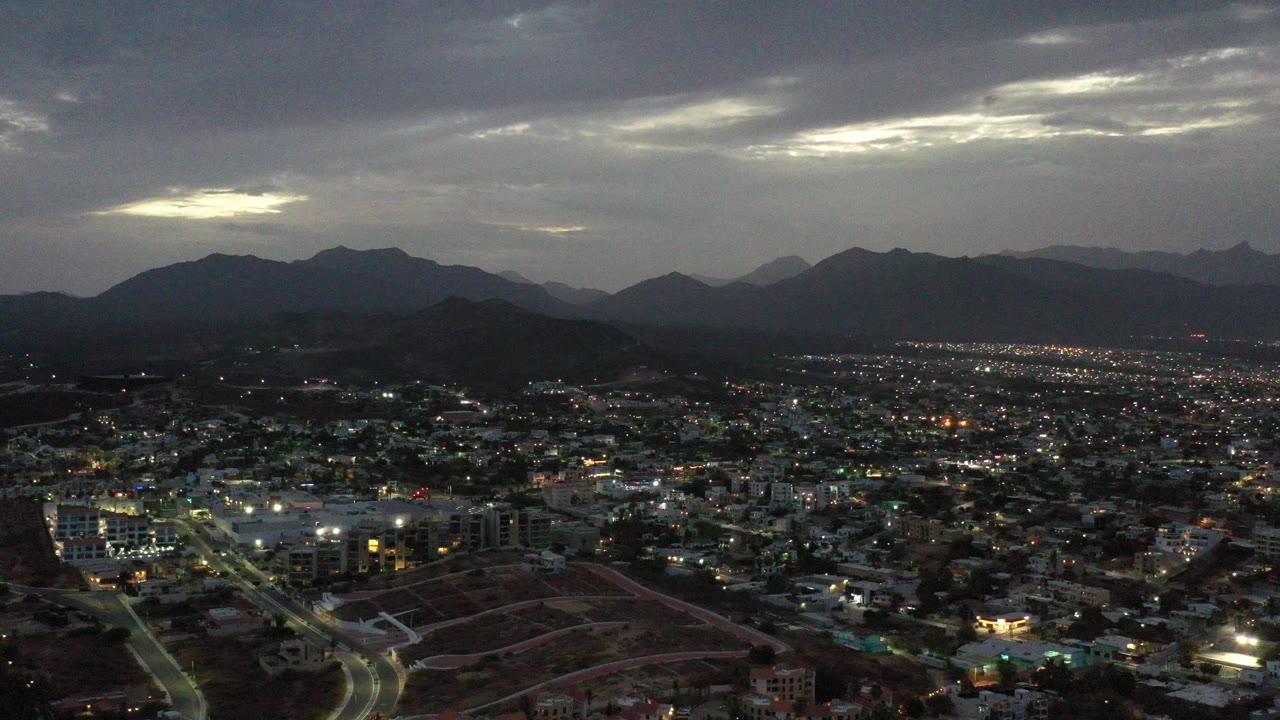 Wide aerial view over Los Cabos, Mexico at dusk, with city lights below dark mountain silhouettes and no visible people.