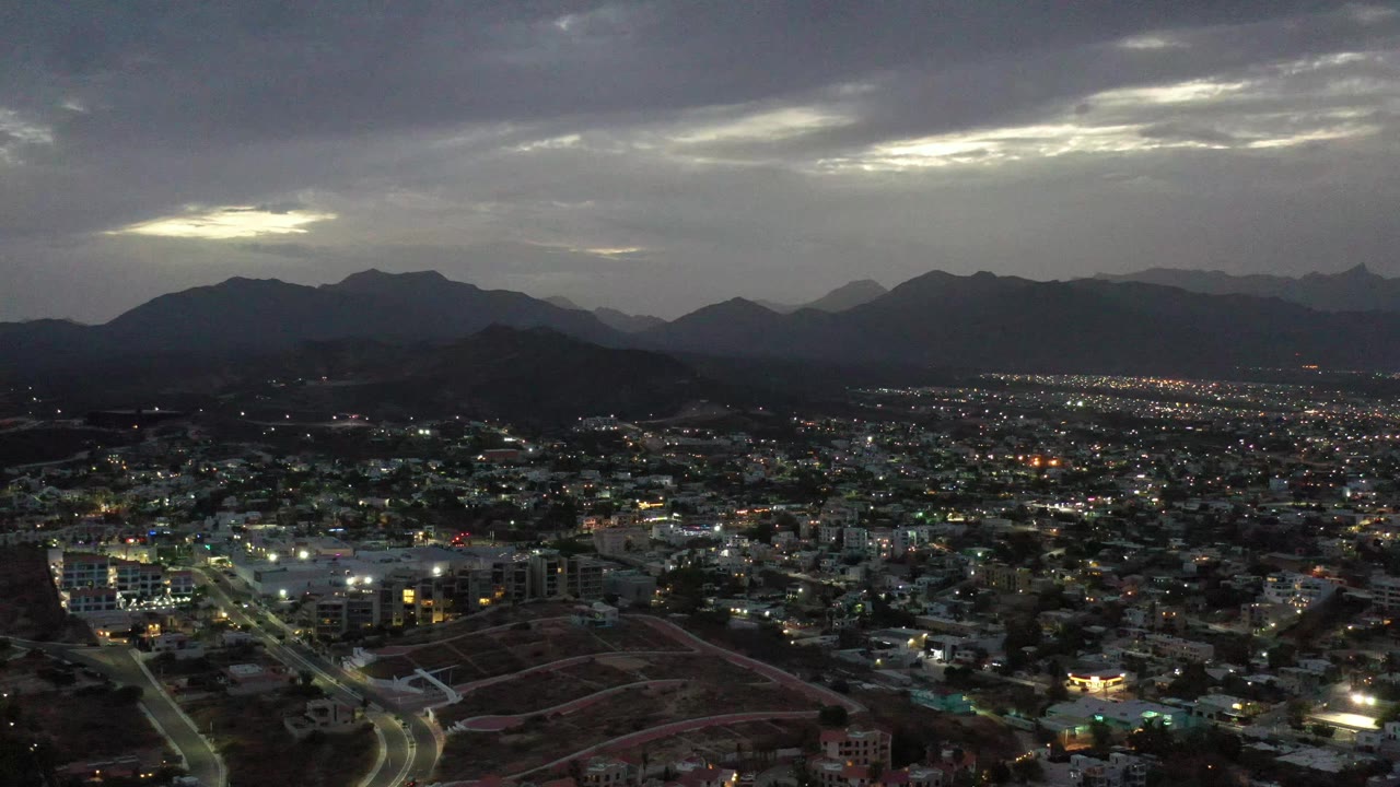 Los Cabos city streets and lit buildings spread beneath a dark mountain ridge and layered gray clouds, with no visible people.