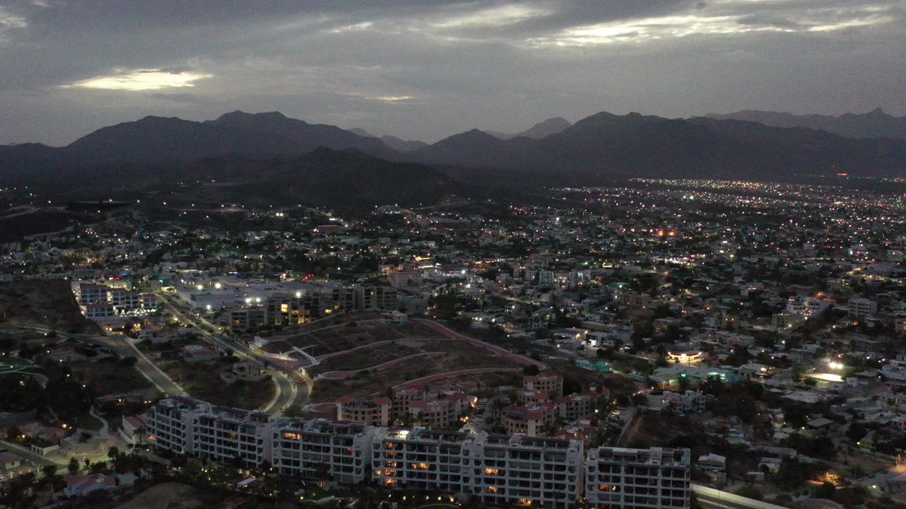 Aerial view over Los Cabos at dusk, with apartment buildings in the foreground, city lights across residential blocks, dark mountains on the horizon, cloudy sky, and no visible people.
