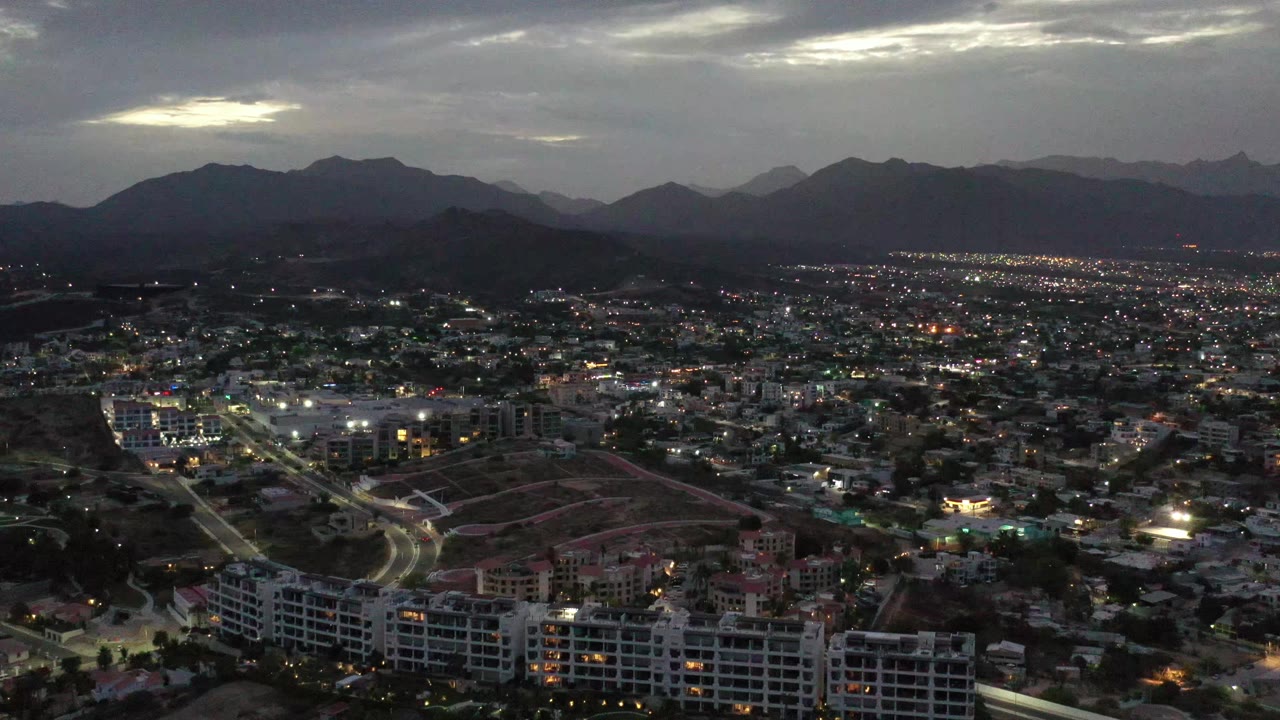 High aerial frame over Los Cabos showing clustered homes, lit roads, foreground apartment blocks, a broad mountain ridge, dim cloud cover, and no visible people.