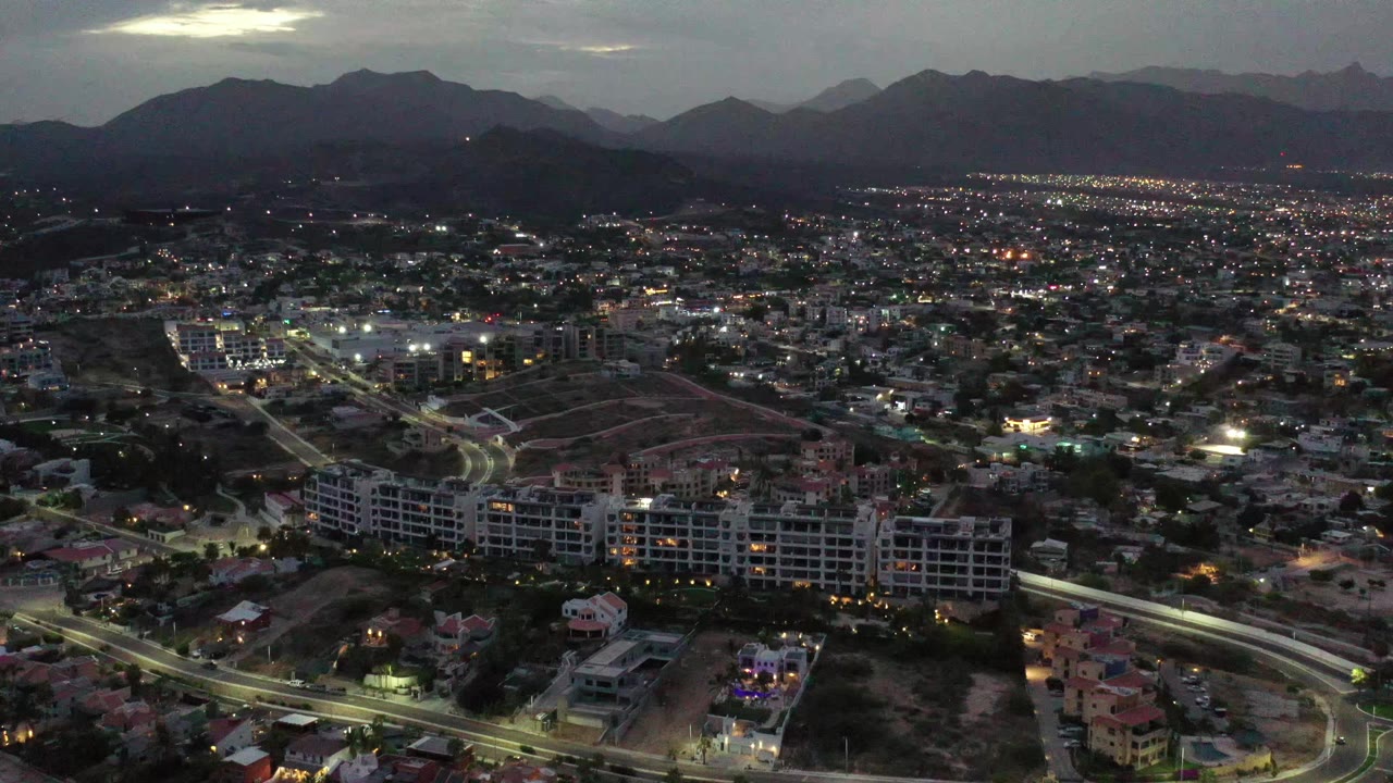 Early aerial frame over Los Cabos, Mexico, with lit roads curving through foreground buildings, a dense urban midground, mountain ridges in back, and no visible people.
