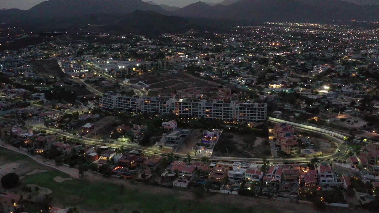 The second frame holds on the Los Cabos neighborhood, with a bright road curve in the lower middle, clustered residences, distant city lights, mountains, and no visible people.