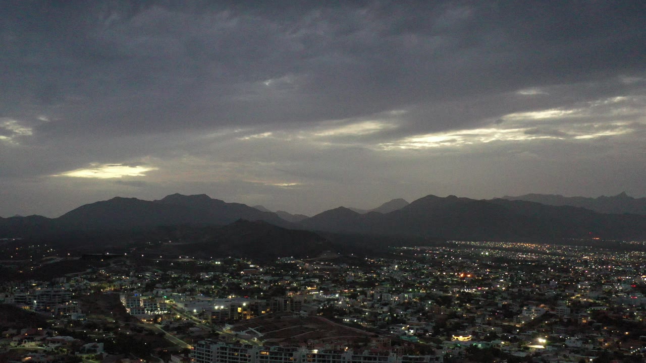 Opening frame of Los Cabos, Mexico, showing a wide spread of city lights below dark mountain silhouettes and a heavy gray cloud ceiling, with no visible people.