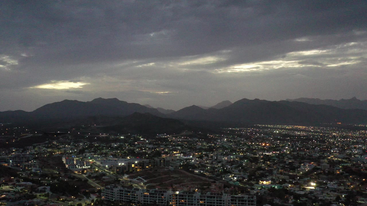 Wide dusk cityscape below dark clouds and mountain silhouettes.