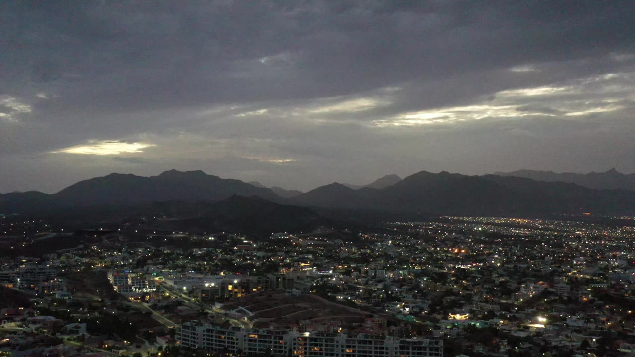Los Cabos city lights spread across the lower frame while a pale break in the gray sunset clouds sits above the dark mountain ridge, with no visible people.