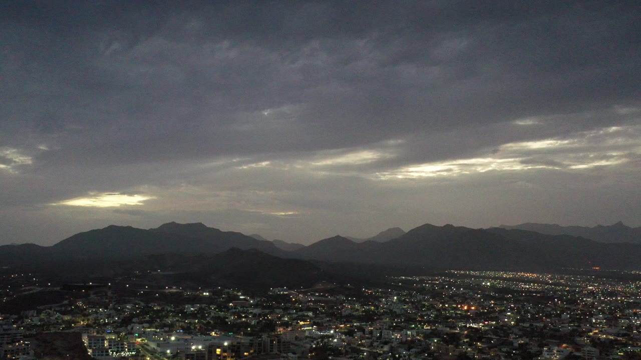 Dusk city lights below dark mountains and a cloudy sky.