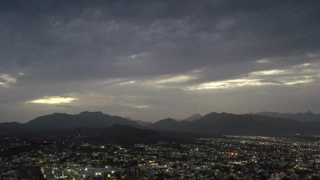 Los Cabos city lights spread across the foreground while a dark ridge line sits under gray clouds and a pale opening of sunset light, with no visible people.