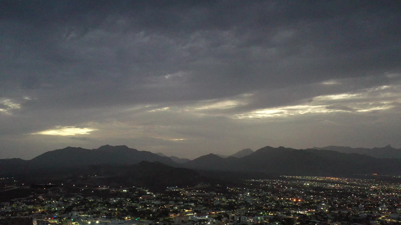 Los Cabos, Mexico city lights spread beneath a dark cloud ceiling and distant mountain silhouettes at the start of the clip, with no visible people.