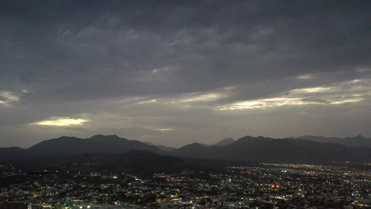 Cloudy sunset sky above city lights and layered mountain silhouettes.