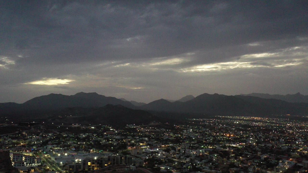 Los Cabos, Mexico city lights spread below dark mountain ridges and a heavy gray sky, with no visible people.