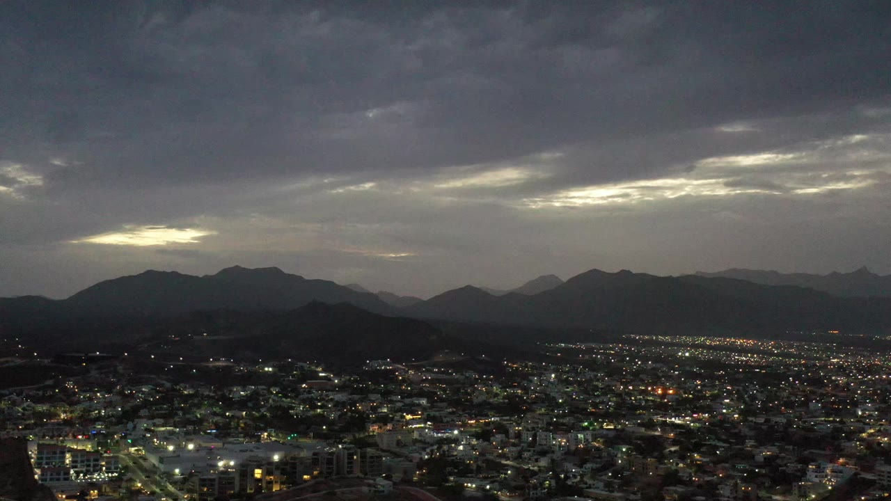 Los Cabos, Mexico appears as a glowing urban grid under layered mountain silhouettes and low gray clouds, with no visible people.