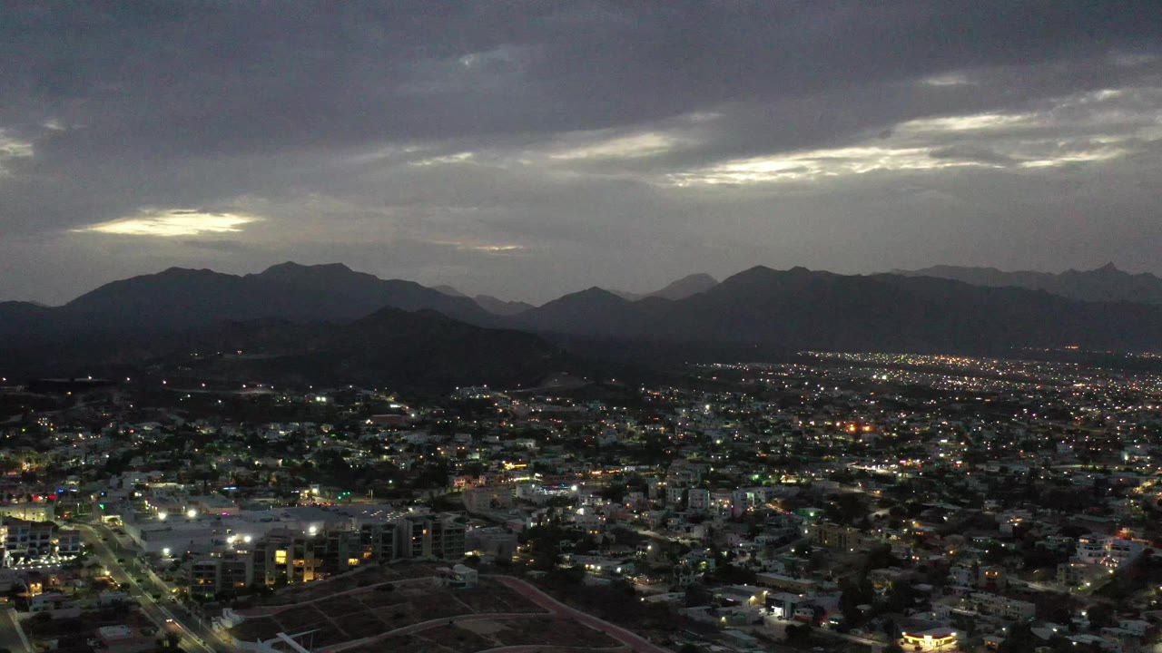 Wide Los Cabos city view from above, with glowing street grids, low dark hills, layered mountain ridges, broken cloud light, and no visible people.