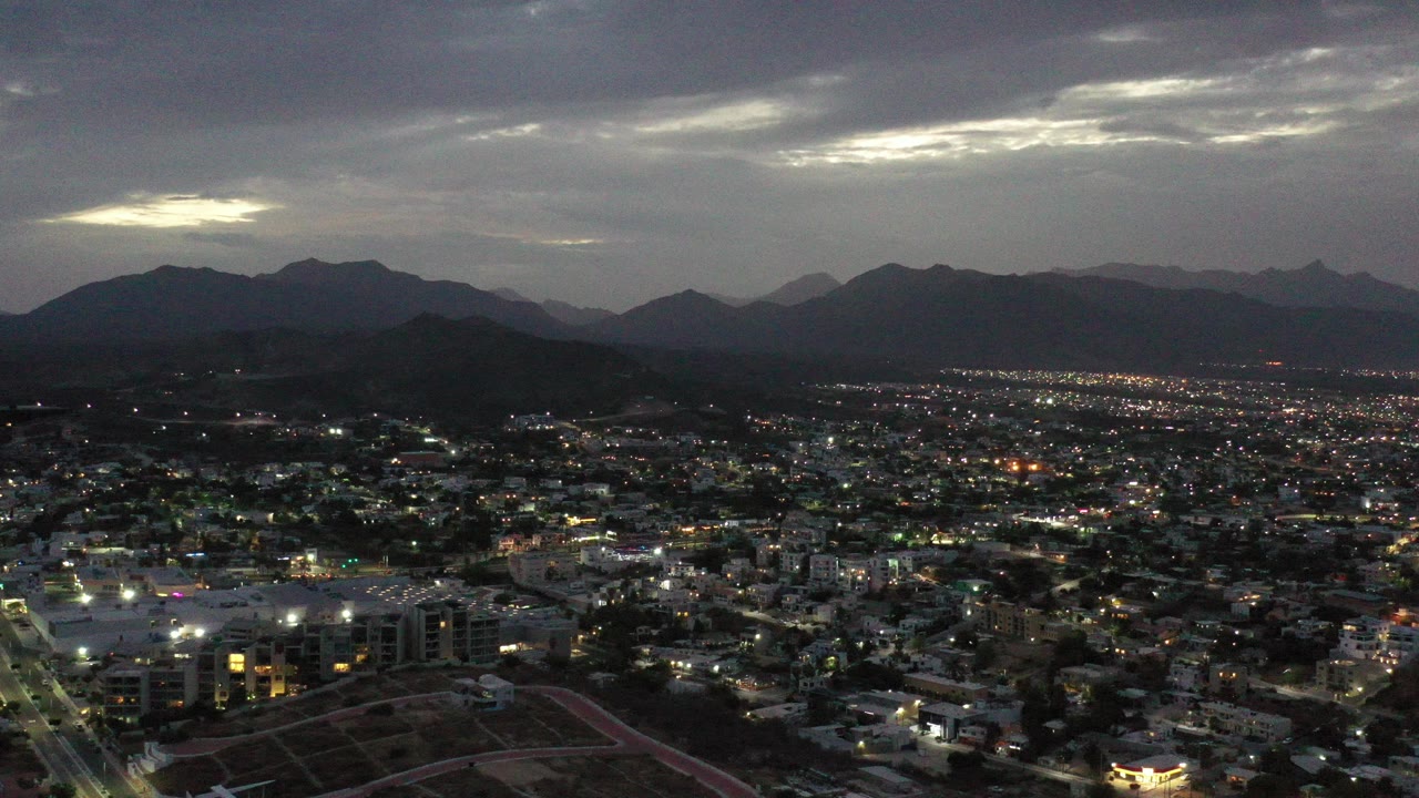 Opening frame of Los Cabos, Mexico from above, with scattered city lights, dark mountain silhouettes, heavy cloud cover, and no visible people.