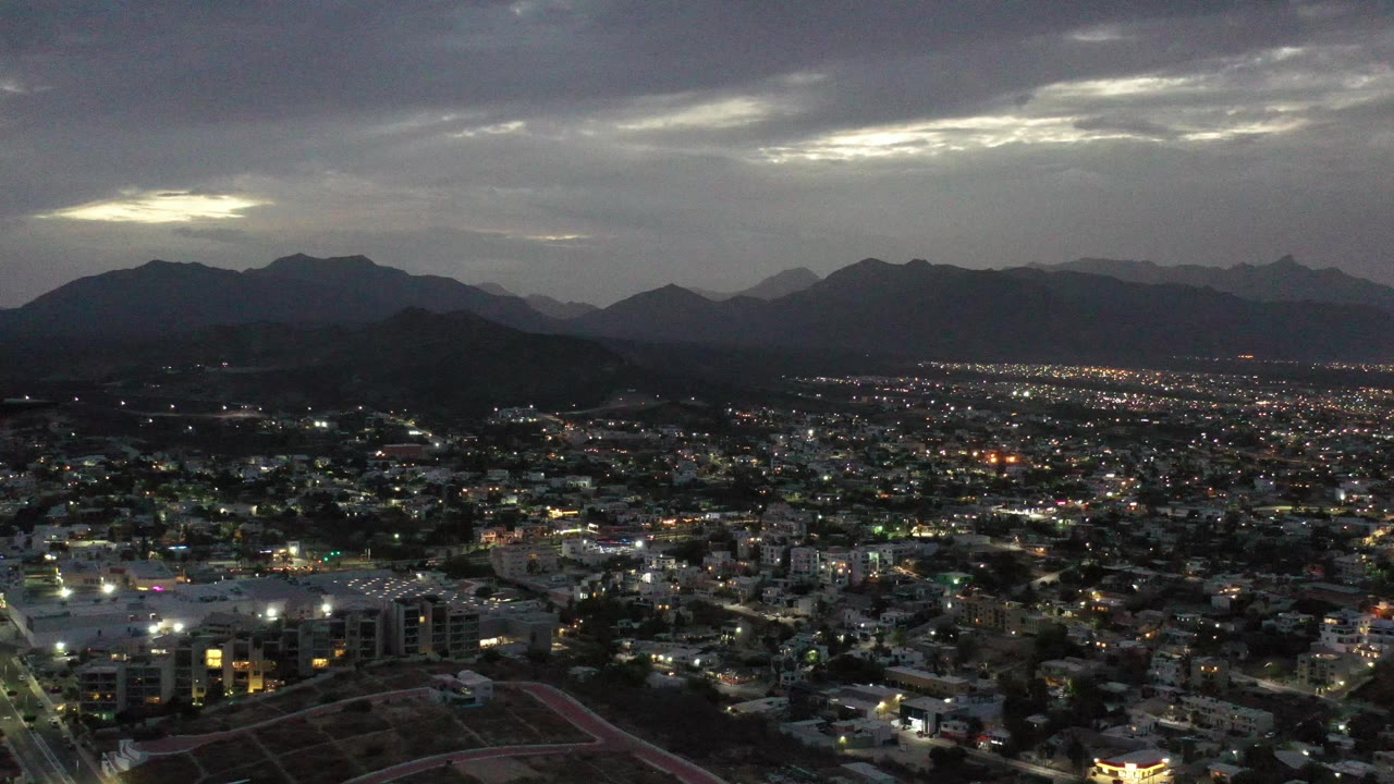 Drone view over city lights with a cloudy sky and mountain backdrop.