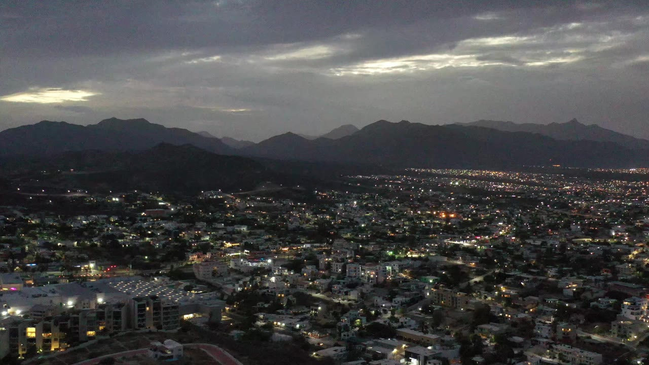 High aerial frame over Los Cabos, Mexico, showing dense urban blocks, illuminated streets, layered mountain silhouettes, and a bright break in the gray cloud cover, no visible people.