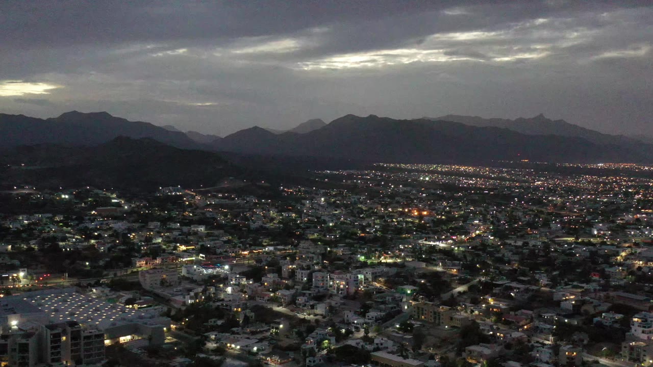 Los Cabos city lights spread across the frame beneath layered mountain ridges and a gray evening cloud deck, with no visible people.