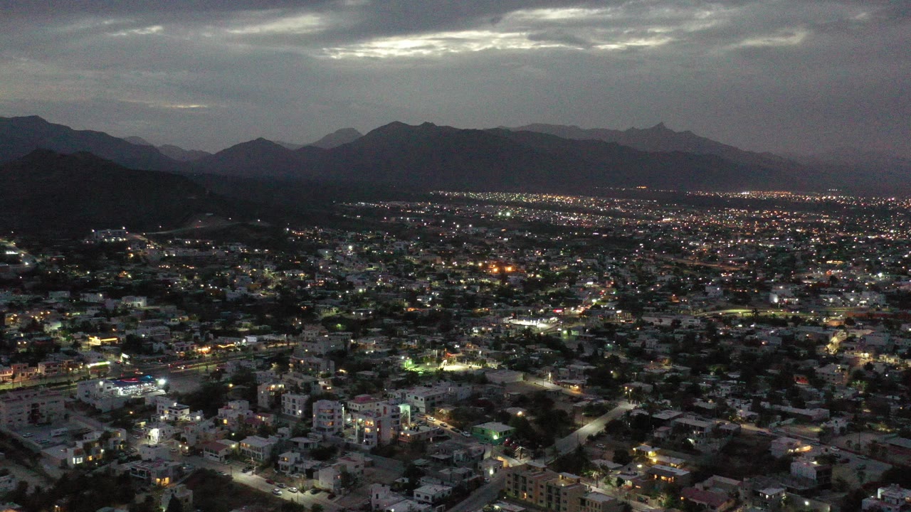 Los Cabos, Mexico night aerial view with lit streets and buildings in the foreground, dark mountain ridges in the distance, cloudy sky above, and no visible people.