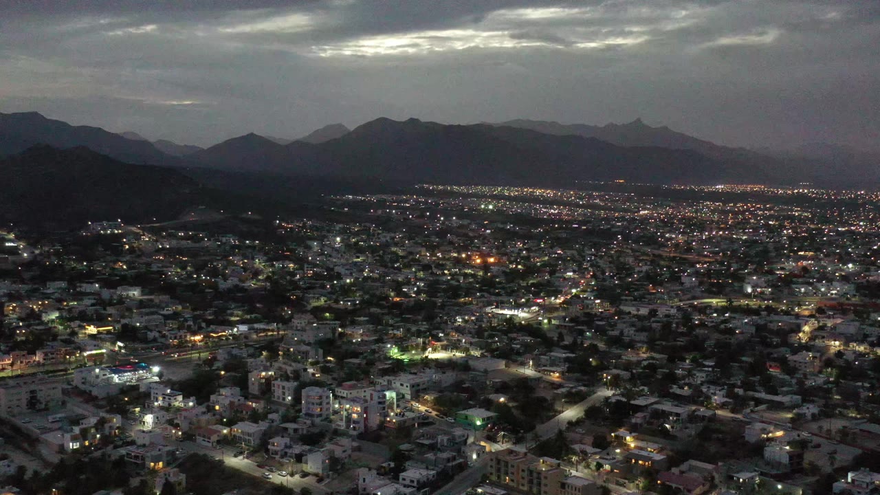 Los Cabos, Mexico appears as a broad night city grid with white and amber street lights, low buildings, dark mountains behind, layered clouds, and no visible people.